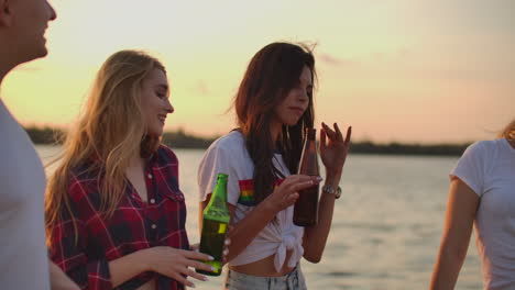 Three-young-girls-with-long-hair-are-dancing-on-the-open-air-party-on-the-beach-with-beer.-They-are-moving-their-young-bodies-on-the-lake-coast-and-having-a-great-time-at-sunset-with-friends.